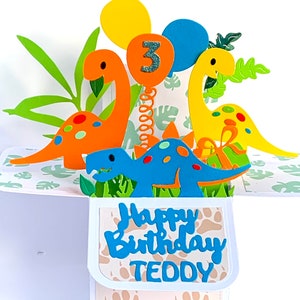 May include: A 3D pop-up birthday card featuring dinosaurs, balloons, and the number 3. The card is decorated with green leaves and a light brown background. The text "Happy Birthday Teddy" is on the front of the card.