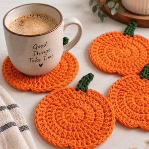 Könnte beinhalten: Ein Keramikbecher mit dem Text Good Things Take Time und einem kleinen Herzsymbol, der auf einem runden, orangefarbenen, gehäkelten Kürbis-Untersetzer mit grünem Stiel steht. Drei weitere Kürbis-Untersetzer liegen daneben auf der Oberfläche.