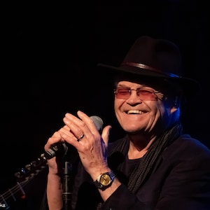 May include: A man wearing a black hat, glasses, and a watch, holding a microphone and smiling. He is wearing a black jacket and a scarf. The background is dark.