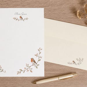 May include: A white stationery sheet featuring the name Olivia Grace in script, decorated with watercolour illustrations of robins perched on leafy branches. An off-white envelope with a matching robin illustration and a cream-coloured ballpoint pen.
