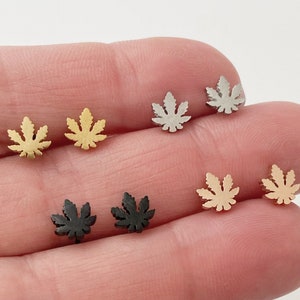 May include: Six pairs of small, silver, gold, and black marijuana leaf stud earrings. The earrings are displayed on a hand.
