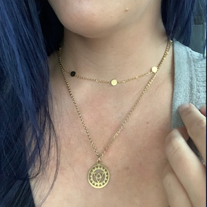 May include: A gold chain necklace with a pendant featuring a geometric design. The pendant is a circle with a triangle inside, surrounded by a ring of smaller triangles. The necklace is layered over a shorter chain with small gold and black beads.