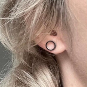May include: A black, circular earring is shown on the ear. The earring has a minimalist design, with a thin, dark outline. The earring is a simple, yet stylish accessory.