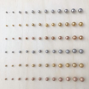 May include: A row of 10 rows of small round beads in various metallic colours, including silver, gold, and rose gold. The beads are arranged in a grid pattern on a light wood surface.