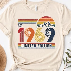 May include: A cream-colored t-shirt featuring a graphic with the text EST. 1969 LIMITED EDITION. The design includes a retro-style sunset, mountain peaks, and pine trees in shades of red, orange, yellow, green, and blue with a distressed texture.