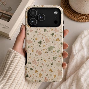 May include: A rectangular smartphone case featuring a cream-colored base with a multi-colored terrazzo pattern of small pink, green, and yellow stone-like flecks. The case has a black camera module cutout with three circular lens openings.