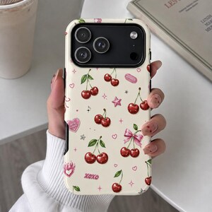 May include: A cream-colored phone case featuring a repeating pattern of red cherries with green stems, pink bows, small pink hearts, stars, and the text 2000s and XOXO. The case has a black camera cutout area with three circular lens openings.