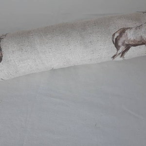 May include: A close-up of a beige fabric with a repeating pattern of brown deer. The fabric is rolled up and the deer are facing the right side of the image.