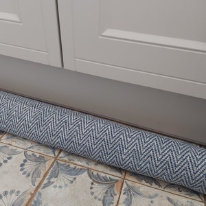 May include: A rolled up blue and white patterned rug with a chevron design. The rug is lying on a tiled floor in front of a white cabinet.