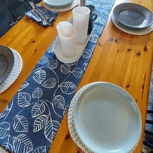 May include: A wooden table with a blue and white leaf patterned table runner. There are four place settings with white plates and woven placemats. Two white candles are on the table runner.