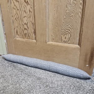 May include: A grey and white patterned doorstop made of fabric. The doorstop is placed on a grey carpet in front of a wooden door.