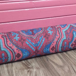 May include: A colourful doorstop with a paisley pattern in shades of pink, blue, and red. The doorstop is made of fabric and is designed to prevent doors from slamming.