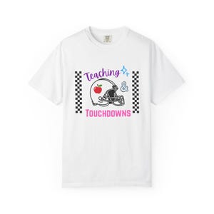 Puede incluir: Una camiseta blanca de manga corta con un gráfico que incluye el texto Teaching & TOUCHDOWNS, un casco de fútbol americano con una manzana roja, una chispa azul y dos bordes verticales a cuadros blancos y negros a los lados.