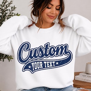 May include: A white long-sleeve crewneck sweatshirt featuring a dark blue distressed graphic design on the chest. The graphic displays the word Custom in a large script font above a banner containing the text YOUR TEXT with a star on each side.