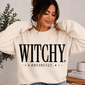 Può includere: Una felpa girocollo color crema con la scritta nera WITCHY, OBVIOUSLY. stampata sul davanti. Il design include piccole stelle a quattro punte ai lati della parola OBVIOUSLY. La felpa presenta maniche lunghe e un colletto a coste.