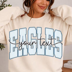 May include: A cream-colored crewneck sweatshirt featuring the word EAGLES in large, blue-outlined block letters with a vertical pinstripe pattern. The words your text are written in a black cursive script across the center of the graphic.