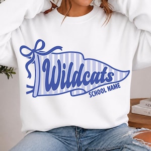 May include: A white long-sleeve crewneck sweatshirt featuring a blue graphic of a pennant flag with vertical stripes. The text "Wildcats" is written in a bold blue script font across the center, with the words "SCHOOL NAME" printed below in blue.