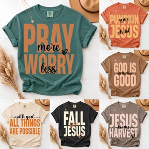 May include: A collection of six short-sleeve crew neck t-shirts in various colors including teal, orange, brown, cream, black, and gray. Each shirt features screen-printed text with religious and seasonal themes, including phrases like PRAY more WORRY less.