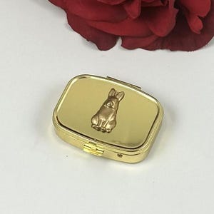 May include: A gold-colored pill box with a rectangular shape and a hinged lid. The top of the box features a raised, bronze-colored bunny rabbit. The box is closed and sits on a white surface, with a red rose in the background.