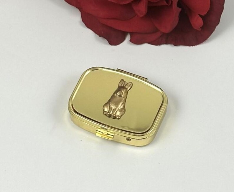 May include: A gold-colored pill box with a rectangular shape and a hinged lid. The top of the box features a raised, bronze-colored bunny rabbit. The box is closed and sits on a white surface, with a red rose in the background.