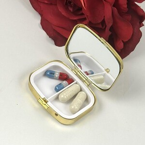 May include: A small, open, gold-colored pill box with a mirror inside. The box contains several capsules in white and red/blue colors. The box is on a white surface, with a red rose in the background.