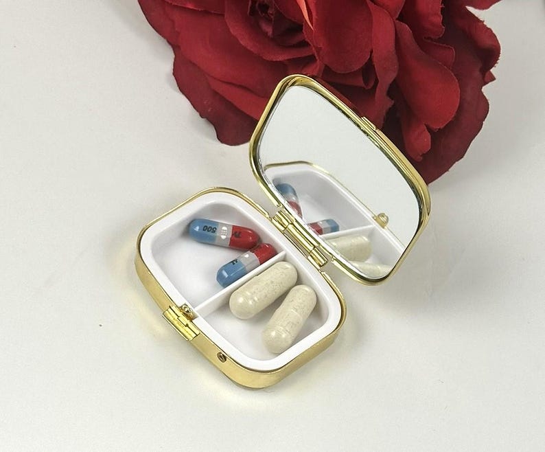 May include: A small, open, gold-colored pill box with a mirror inside. The box contains several capsules in white and red/blue colors. The box is on a white surface, with a red rose in the background.
