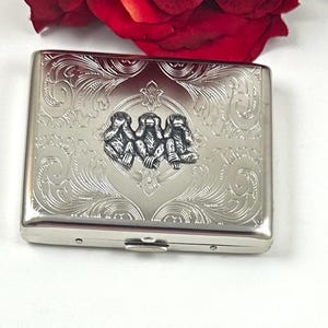May include: A rectangular silver-toned metal cigarette case featuring an embossed design of the three wise monkeys in the center. The surface is decorated with intricate scrollwork and floral patterns surrounding the central monkey motif.