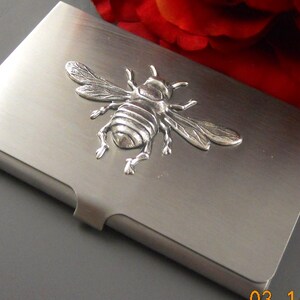 K&ouml;nnte beinhalten: Ein silberner Metall-Visitenkartenhalter mit einem 3D-Bienendesign.