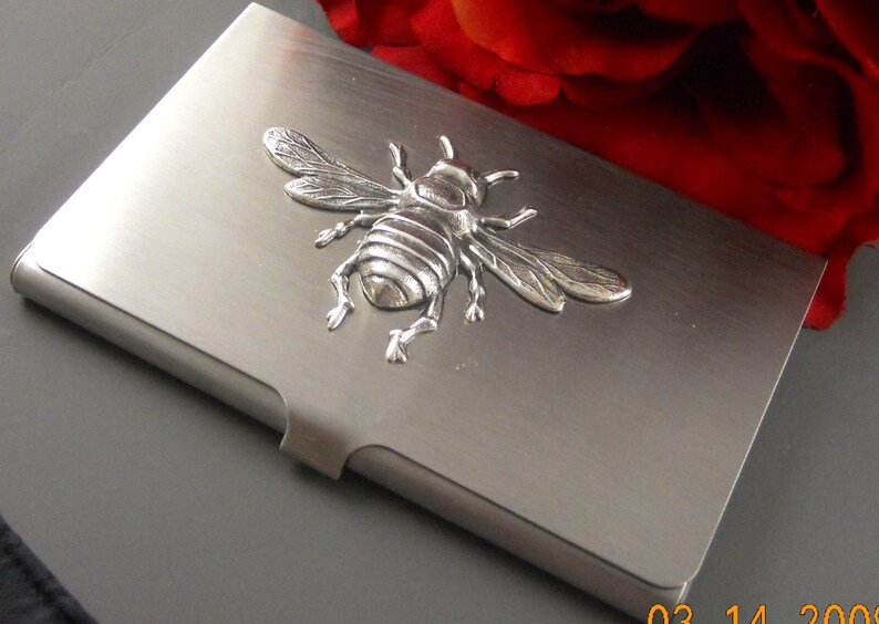 K&ouml;nnte beinhalten: Ein silberner Metall-Visitenkartenhalter mit einem 3D-Bienendesign.