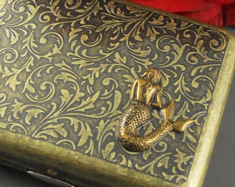 May include: A gold metal cigarette case with a mermaid design. The case has a floral pattern embossed on the surface.