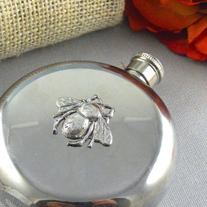 May include: A silver flask with a bee design on the front. The flask is round and has a silver cap.