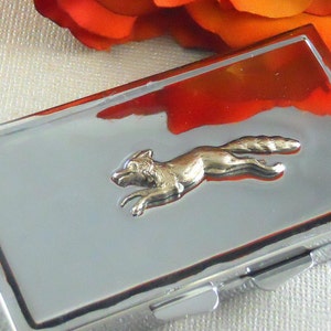 May include: A silver metal pill box with a gold fox design. The fox is running and has a raised design.