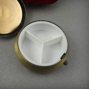 May include: A round, antique gold pill box with a hinged lid and three compartments inside. The box is open, revealing the white interior.