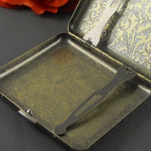 Puede incluir: Un estuche de cigarrillos de lat&oacute;n antiguo con un patr&oacute;n floral en el interior. El estuche tiene una tapa con resorte y un porta cigarrillos incorporado.
