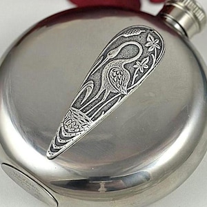 Könnte beinhalten: Eine silberne Trinkflasche mit einem gravierten Design aus einem Kranich und floralen Elementen. Die Flasche hat eine runde Form und einen Schraubverschluss.