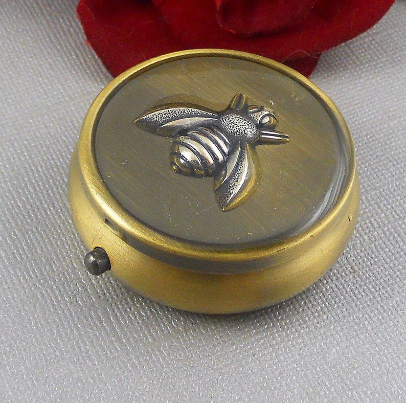 May include: A small, round, gold-colored metal trinket box with a hinged lid. The lid features a raised, silver-colored bee design.