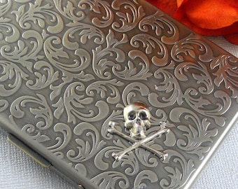 May include: A silver cigarette case with an ornate floral design and a skull and crossbones emblem.