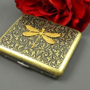 Puede incluir: Una cajetilla de cigarrillos de metal dorado con un dise&ntilde;o floral negro y dorado y una lib&eacute;lula dorada en la parte delantera.
