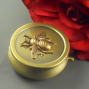 May include: A gold round metal container with a bee design on the lid. The bee is made of gold metal and has a textured surface.