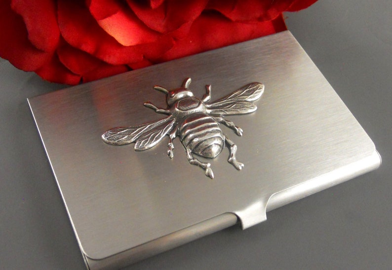 K&ouml;nnte beinhalten: Ein silberner Metall-Visitenkartenhalter mit einem 3D-Bienendesign.