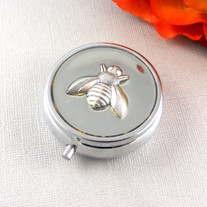 May include: A silver pill box with a bee design on the lid. The bee is made of gold-colored metal and has a textured surface.