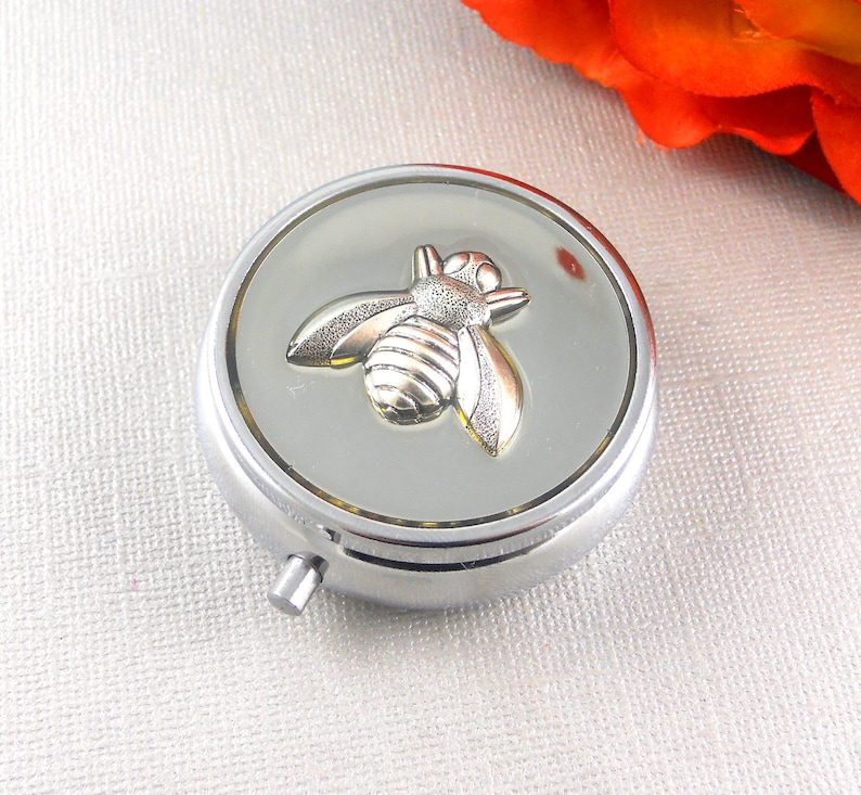 May include: A silver pill box with a bee design on the lid. The bee is made of gold-colored metal and has a textured surface.