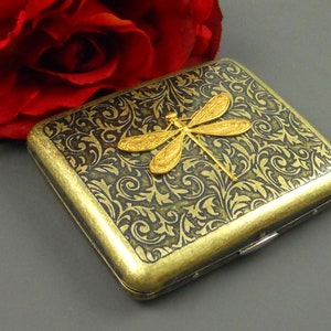 Puede incluir: Una cajetilla de cigarrillos de metal dorado con un patr&oacute;n floral negro y dorado y un dise&ntilde;o de lib&eacute;lula dorada.