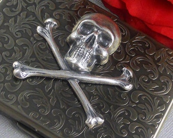 May include: A silver skull and crossbones design on a black metal cigarette case with a floral pattern.