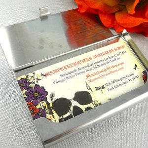 K&ouml;nnte beinhalten: Ein silberner Metall-Visitenkartenhalter mit einer Karte darin. Die Karte hat ein florales Design mit violetten Blumen und einer Totenkopfgrafik. Der Text auf der Karte lautet "Hanocelebrates/Maniadesign", "Steampunk Accessories Jewelry Lockets Cuff links", "Vintage Retro Future Inspired Romantic Lockets", "Maniadesign@etsy.com", "Manocelebrates@etsy.com", "3226 Whooping Crane", "Run Kissimmee Fl 34741".