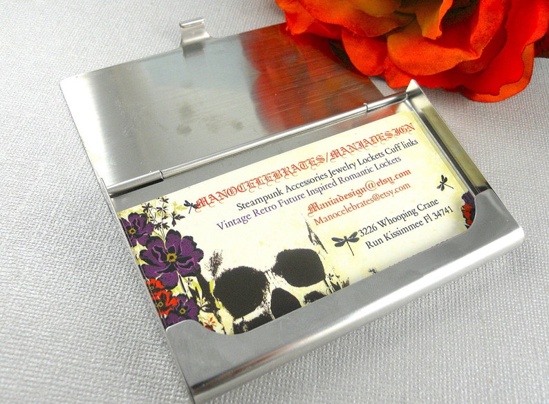 K&ouml;nnte beinhalten: Ein silberner Metall-Visitenkartenhalter mit einer Karte darin. Die Karte hat ein florales Design mit violetten Blumen und einer Totenkopfgrafik. Der Text auf der Karte lautet "Hanocelebrates/Maniadesign", "Steampunk Accessories Jewelry Lockets Cuff links", "Vintage Retro Future Inspired Romantic Lockets", "Maniadesign@etsy.com", "Manocelebrates@etsy.com", "3226 Whooping Crane", "Run Kissimmee Fl 34741".