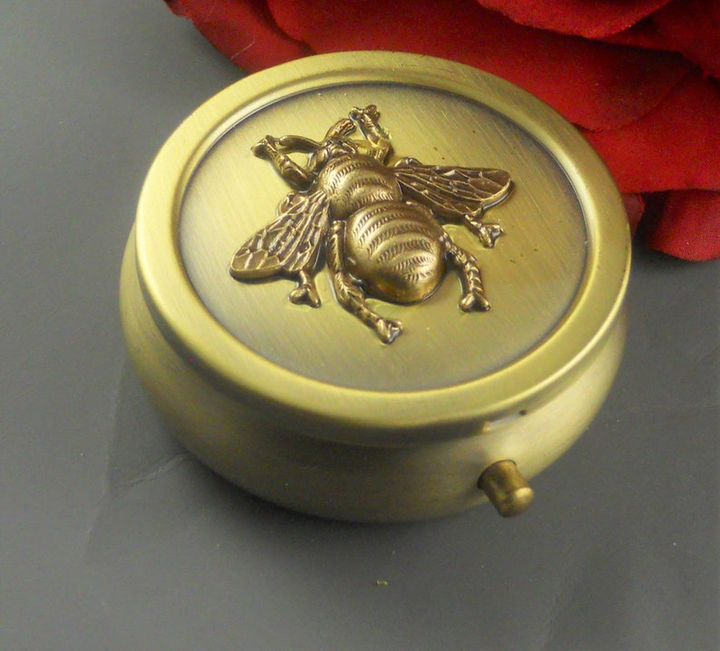 May include: A round, brass trinket box with a hinged lid. The lid features a raised, gold-colored bee design.