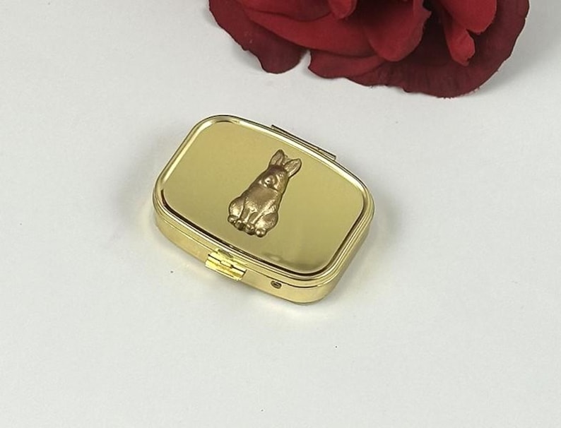 May include: A gold-colored rectangular pill box with a raised bunny design on the lid. The box is closed and has a small clasp. A dark red rose is in the background.