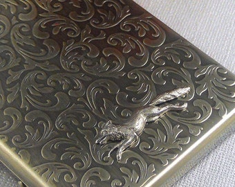 May include: A gold metal cigarette case with an ornate, engraved floral design. A silver fox figurine is attached to the case.