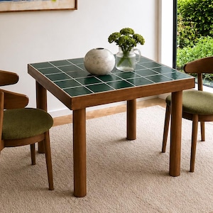 Pode incluir: Uma mesa de jantar quadrada com tampo de azulejos verde-escuros e estrutura de madeira, acompanhada por duas cadeiras de madeira com encostos curvos e assentos estofados em verde-oliva. Uma esfera de mármore branco e um vaso de vidro com flores.